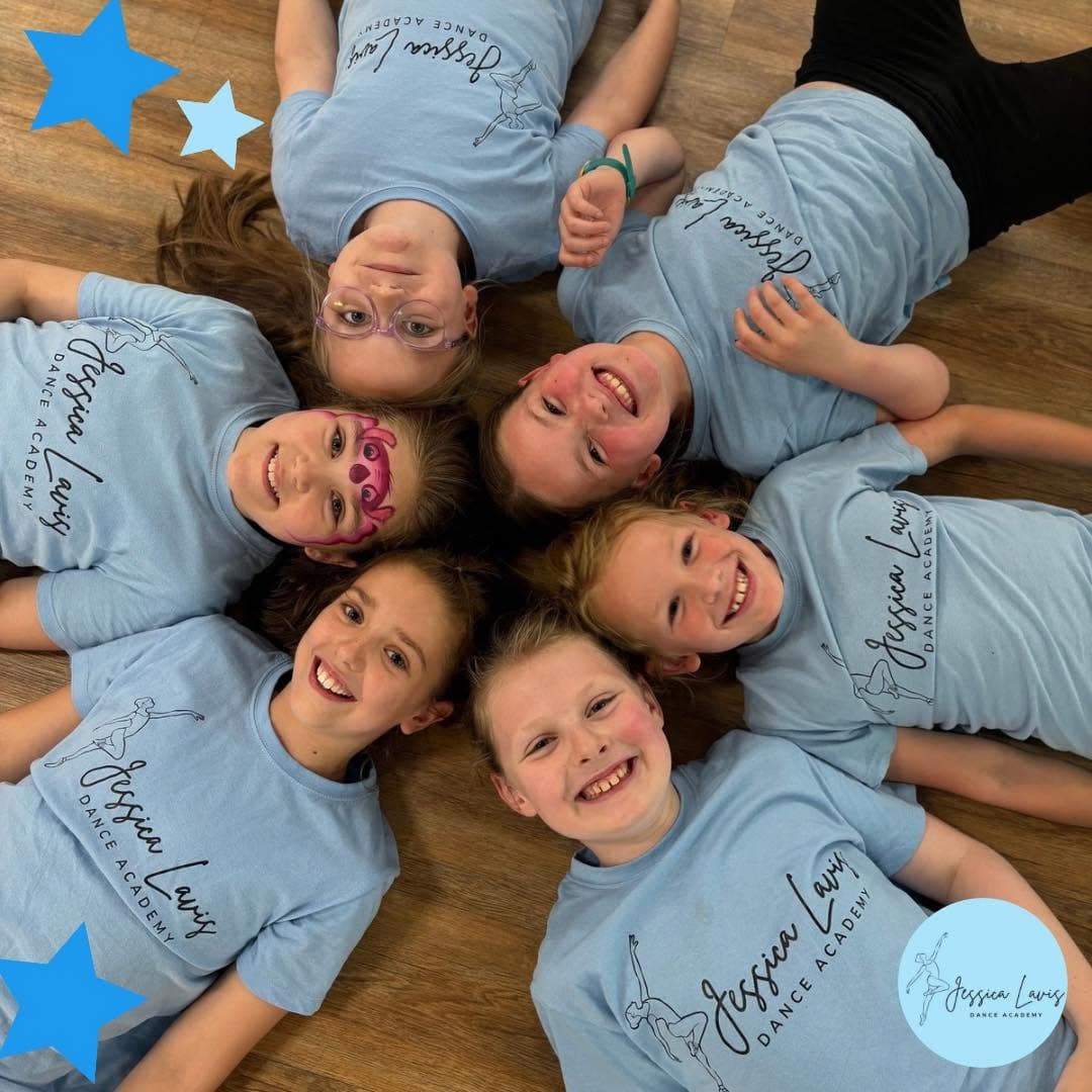 Six smiling dancers lying in a circle on the studio floor in academy t-shirts