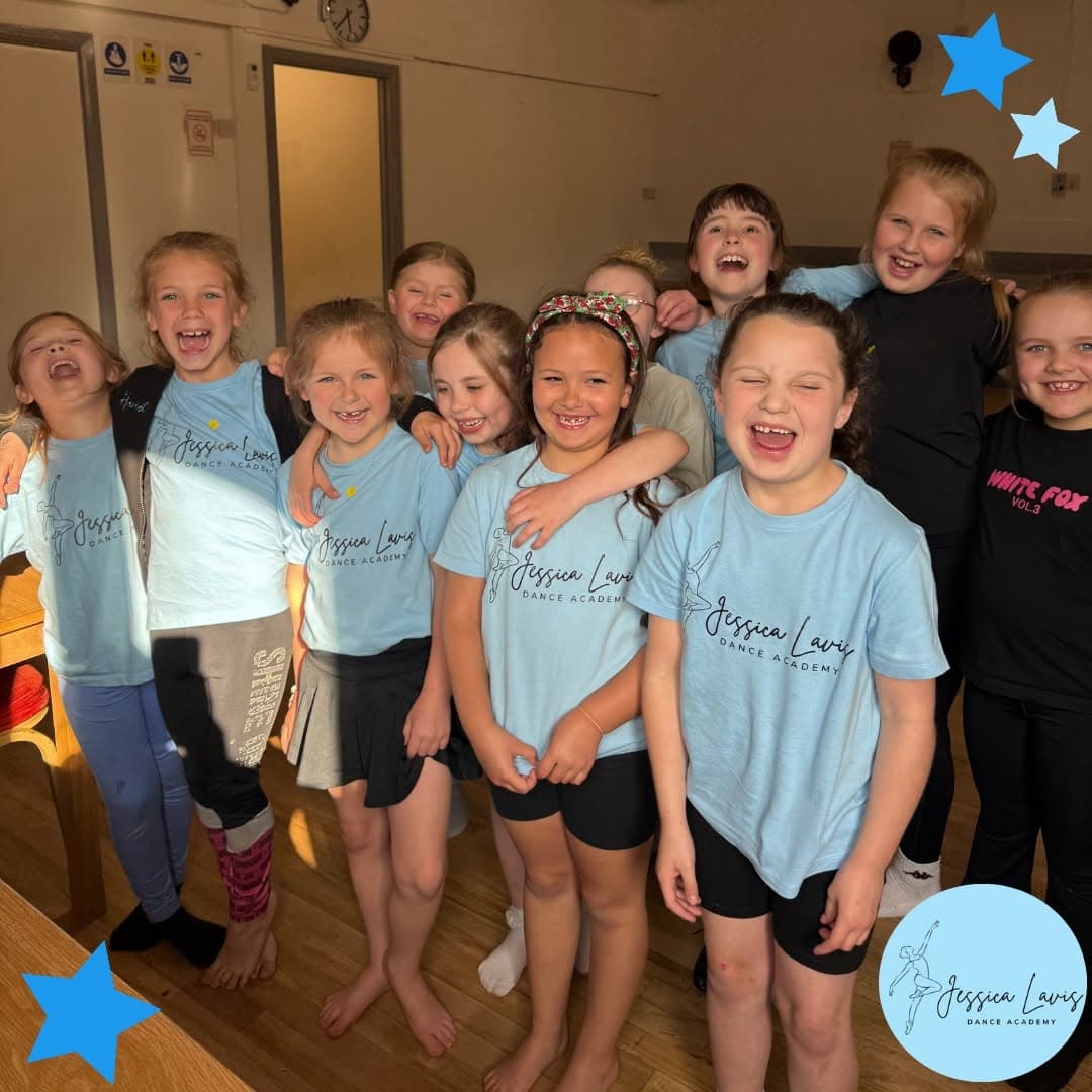 A group of young dancers in light blue academy t-shirts laughing together in the studio