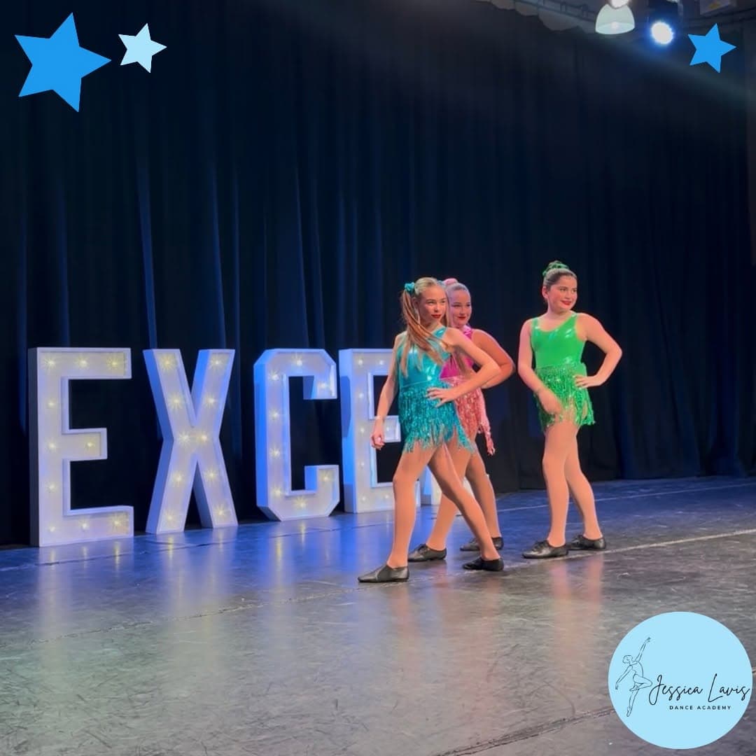 Three dancers in shiny fringed costumes performing beside giant illuminated EXCEL letters
