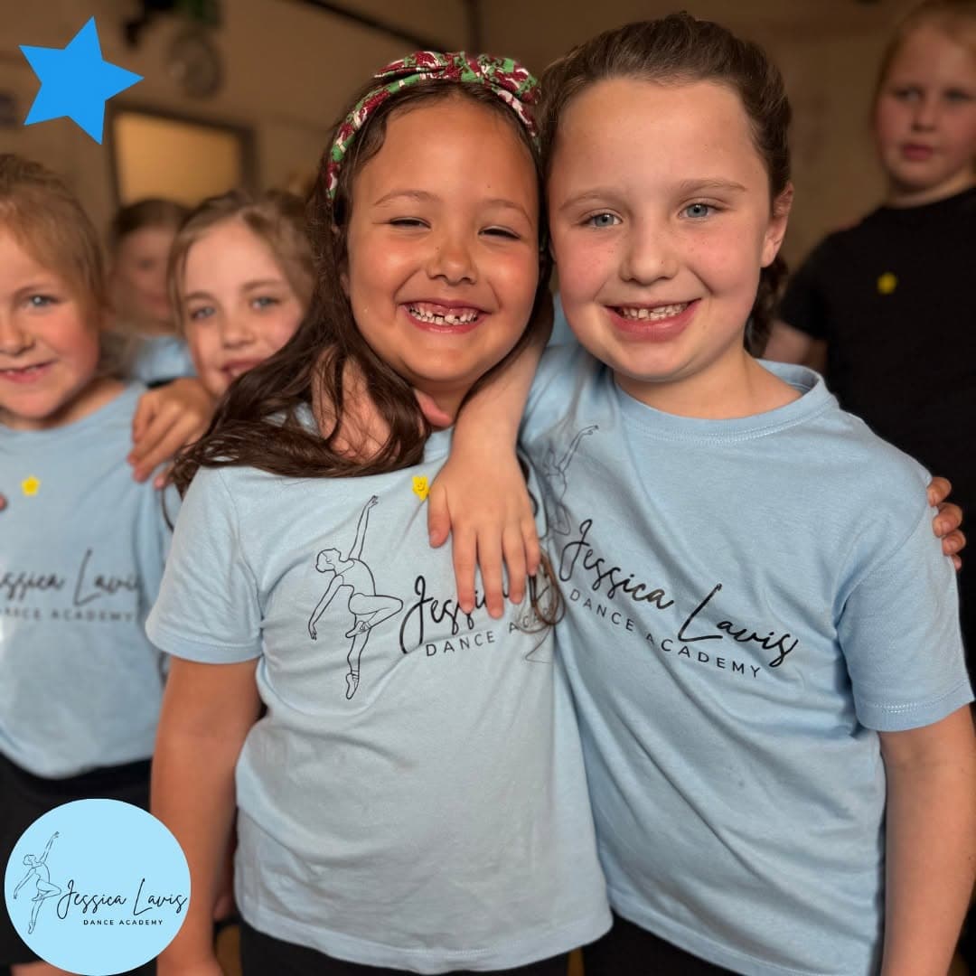 Two young dancers in light blue academy t-shirts hugging and smiling in class