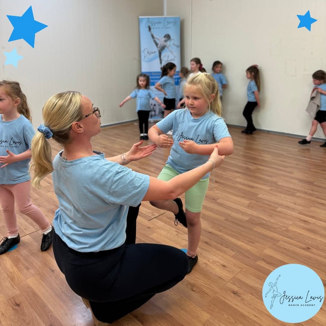 Miss Jess kneeling to help a young dancer balance during a minis class